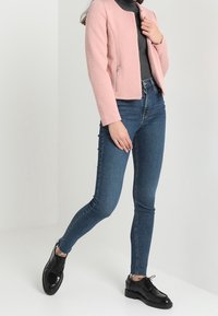 Veste courte rose texturée, col roulé gris moulant, jeans skinny bleus, et chaussures noires à lacets. Design minimaliste avec des fermetures éclair.