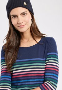 Beanie blu navy con un'emblema di un'ancora gialla. Un top a maniche lunghe presenta strisce orizzontali in rosa, verde, blu e bianco su uno sfondo blu navy.
