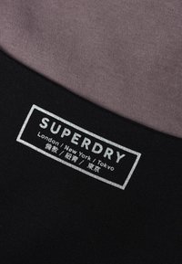 Svart tyg med en grå etikett som visar "SUPERDRY" och stadens namn: London, New York, Tokyo. Slät textur, rektangulär etikettform.