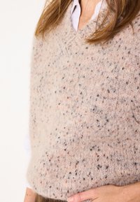 Veste en tricot beige avec col en V, présentant une surface texturée et des taches de tons marron, gris et pêche. Matériau doux et chaud.