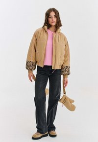 Žena v béžovom pufri s leopardími rukávmi, ružovom svetri, čiernych džínsoch a béžových čižmách, ktorá drží zodpovedajúce béžové rukavice za popruhy.