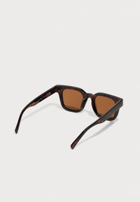 Lunettes de soleil en écaille de tortue avec des verres marron. Design de monture angulaire et épaisse avec une texture brillante. Branches larges avec des détails subtils.