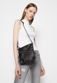 Černá kožená crossbody taška s texturovaným povrchem, zipovým oddílem a nastavitelným popruhem, nošena na bílém pozadí.