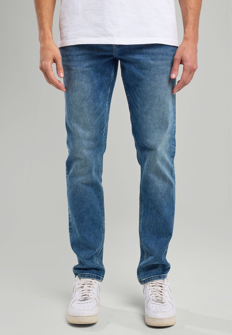 Blue denim jeans with a slim fit, featuring a faded wash, front pockets, and white sneakers. Smooth texture and traditional five-pocket design.