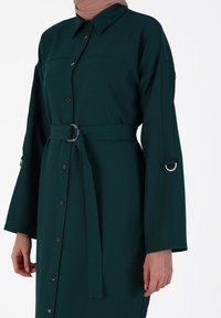 Long, button-up dress in dark green fabric with a collar, belted waist, and wide sleeves featuring metal accents. Smooth texture.