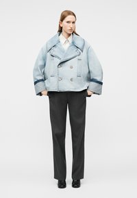 Frau trägt eine übergroße hellblaue Jeansjacke, weißes Hemd mit Kragen, dunkelgraue Hose und schwarze Schuhe, steht mit den Händen in den Taschen.