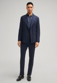Veste de costume bleu marine à double boutonnage avec un pantalon assorti ; présente des revers pointus, des poches avant et des chaussures noires élégantes, créant un look cohérent.