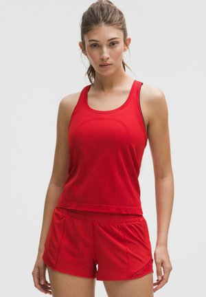 Mladá žena v červenom tielku bez rukávov a červených športových šortkách na svetlom jednoduchom pozadí.