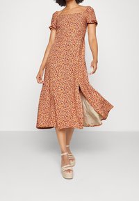 Robe midi fleurie marron avec des manches courtes à volants, un décolleté carré et une fente sur le côté. Portée avec des sandales à plateforme de couleur claire.