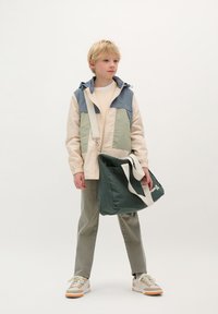 Veste légère multicolore avec capuche ; sac vert et beige avec une bandoulière blanche ; pantalon beige ; baskets avec des accents crème et orange.