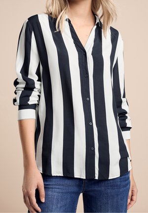 Kvinde iført sort og hvidt lodret striped langærmet skjorte med blå jeans mod beige baggrund.