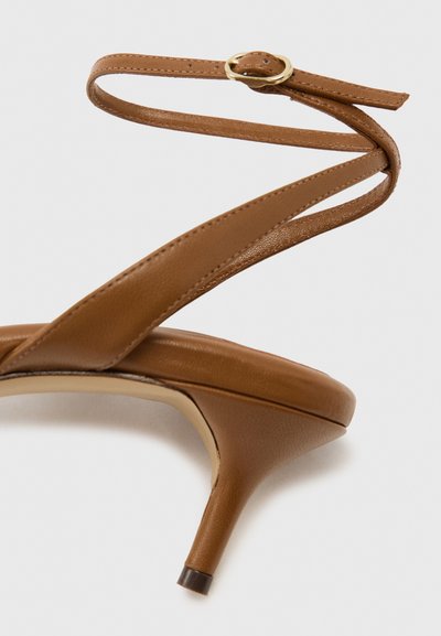 Καφέ δερμάτινο σανδάλι με ψηλό τακούνι, λεπτό λουράκι στον αστράγαλο και χρυσή αγκράφα, μυτερή άκρη τακουνιού και επενδεδυμένη σόλα σε ανοιχτό φόντο.