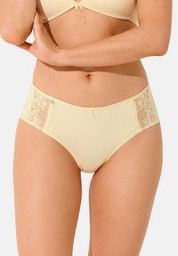 Sous-vêtements beige avec des panneaux latéraux en dentelle. Tissu lisse avec une texture douce. Présente un petit détail en forme de nœud à la taille. Conception taille mi-haute.
