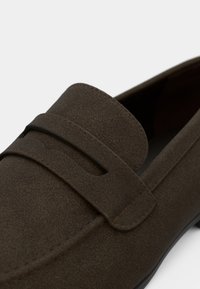 Mocassin en cuir marron avec une surface lisse et texturée, doté d'un bout arrondi et d'une bride décorative traversant le dessus.