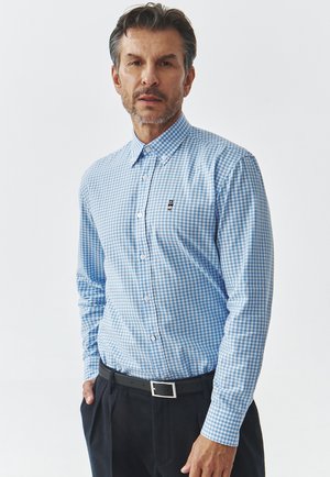 Camisa de botones a cuadros en azul claro y blanco con cuello, que presenta un pequeño logo en el pecho y mangas largas, combinada con pantalones oscuros.