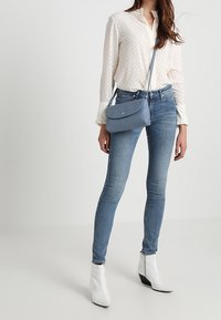 Borsa a tracolla in pelle azzurra chiaro con chiusura a patta, texture liscia e tracolla sottile. Abbinata a una camicetta bianca testurizzata e jeans blu.