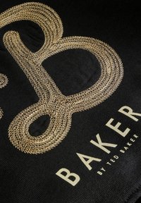Sort stof med tekstureret guld broderet "B" over ordet "BAKER" og mindre tekst "BY TED BAKER" trykt nedenunder.