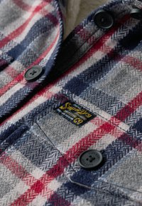 Flanelskjorte med et mønster i blå, rød og grå firkanter, sorte knapper og et mærke med teksten "Superdry" på en tekstureret overflade.
