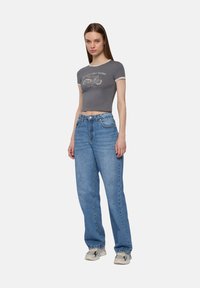 Młoda kobieta stoi w zrelaksowanej pozycji, ubrana w szary, krótki T-shirt z nadrukiem motocykla, jeansy o wysokim stanie i sneakersy.