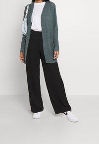 Dlouhý kardigan s otevřeným předním dílem v texturovaném teal pletení, spárovaný s bílým tričkem a černými širokými kalhotami. Bílé tenisky a světle modrá taška.