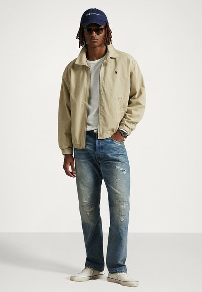Giacca bomber beige, t-shirt bianca, jeans blu strappati, sneakers bianche, cappellino da baseball blu navy e occhiali da sole rotondi.