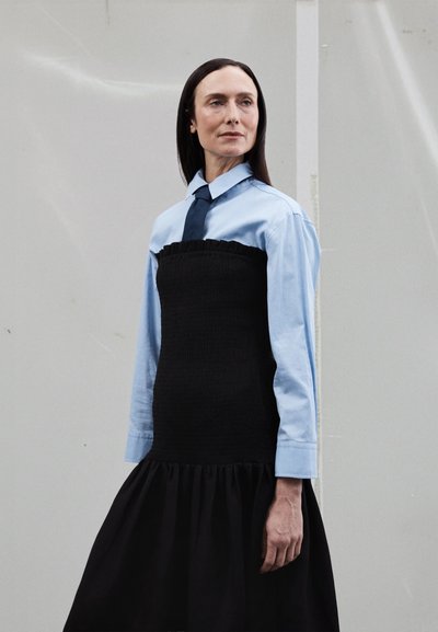 Fekete smock ruha fodros felsőrésszel, világoskék gombos ing fölött rétegezve, sötét nyakkendővel, bővülő szegéllyel. Simaság, hétköznapi design.