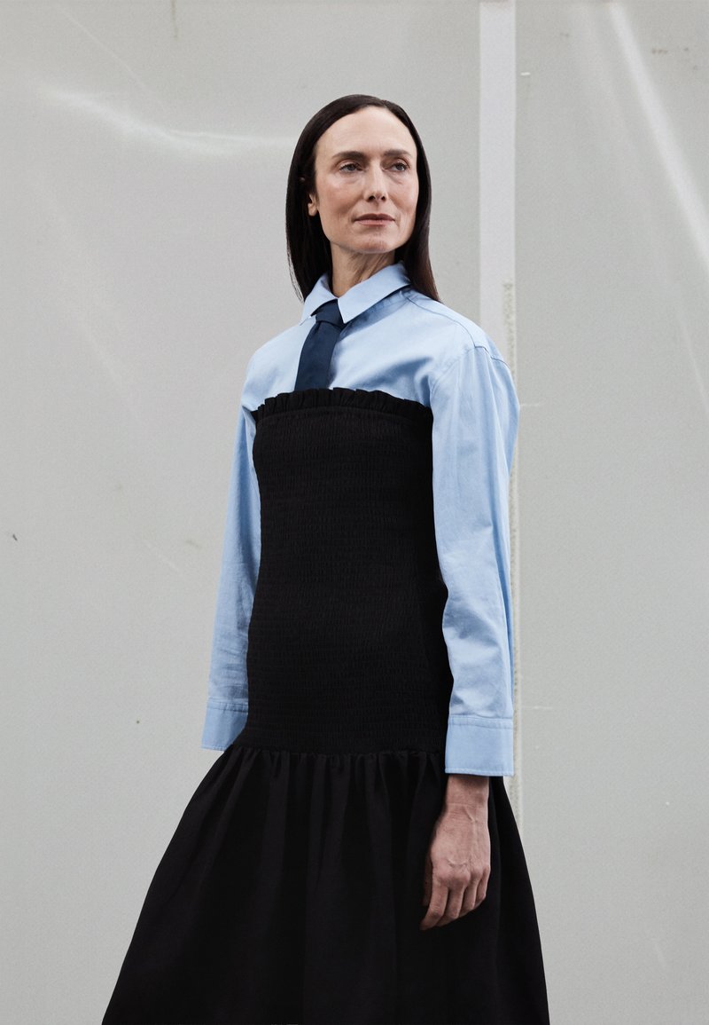 Juoda raukšlėta suknelė su raukšlėtu viršumi, dėvima ant šviesiai mėlynos palaidinės su sagutėmis ir tamsiai mėlynos kaklaraiščio, su plačiomis apačiomis. Lygios tekstūros, kasdienis dizainas.