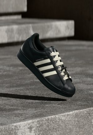Zwarte leren sneaker met witte strepen en veters, voorzien van een rubberen neus en een getextureerde zool. "SUPERSTAR" is op de zijkant gegraveerd.