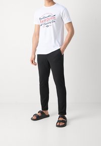 Biała bawełniana koszulka z graficznym nadrukiem, w połączeniu z czarnymi spodniami slim-fit oraz czarnymi sandałami z regulowanymi paskami i teksturowaną podeszwą.