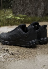Scarpe da trekking nere con tessuto Gore-Tex e suole robuste posizionate su un sentiero esterno fangoso con erba e corteccia d'albero sullo sfondo.