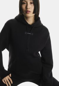 Schwarzer Hoodie aus weichem, strukturiertem Stoff. Verfügt über eine frontale Kängurutasche, Zugbänder und ein Logopatch auf der Brust. Einfaches Design.