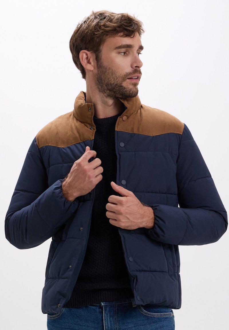 Giacca puffer da uomo in blu navy con spalle in suede marrone, chiusure a scatto e texture trapuntata. Presenta un colletto alto e polsi elastici.