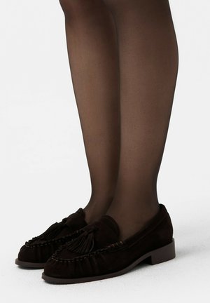 Mocassini in suede marrone scuro con dettagli a nappina, tacco piatto e accentuazioni cucite, indossati con collant neri trasparenti.