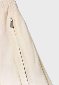 Tissu beige avec détail de poche à fermeture éclair verticale en métal et taille élastiquée froncée sur un vêtement léger.