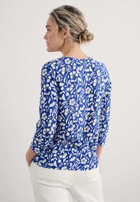 Langärmliges Oberteil mit blauem Blumenmuster und rundem Ausschnitt. Es zeichnet sich durch einen strukturierten Stoff und weiße Blumenakzente auf einem blauen Hintergrund aus.