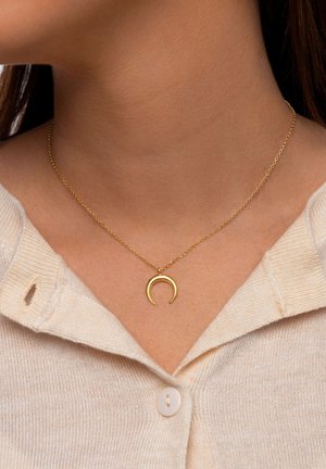 Nahaufnahme einer Person, die eine goldene Halskette mit einem Halbmondanhänger an einer dünnen Kette trägt, kombiniert mit einem beigen, geknöpften Pullover.