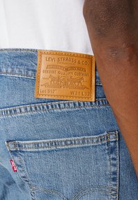 Blå denimjeans med en kvadratisk läderpatch i tanfärg som har "Levi Strauss & Co."-logotyp, sömnad på bakfickan och en röd etikett.
