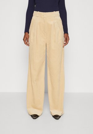 Kobieta ubrana w beżowe szerokie spodnie sztruksowe z plisami i czarną bluzkę z długim rękawem, stojąca na tle jasnego, jednolitego tła.