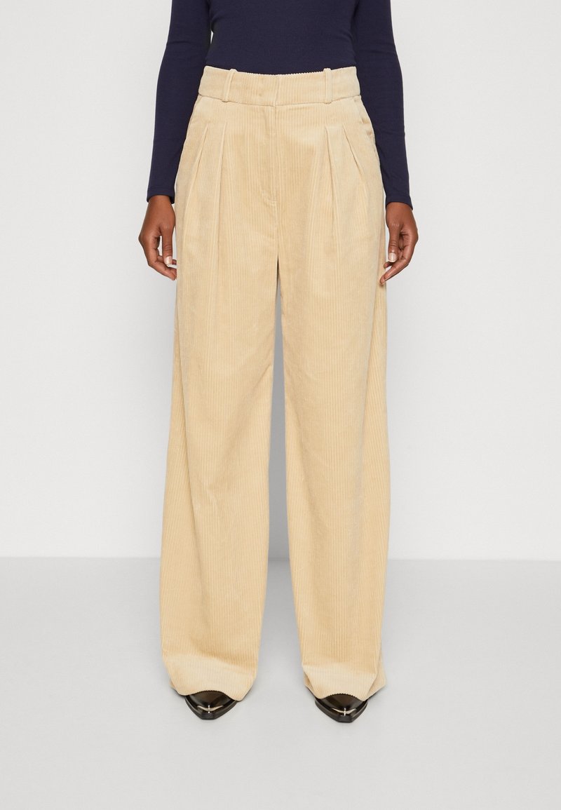 Femme portant un pantalon large en velours côtelé beige avec des plis et un haut noir à manches longues, debout devant un fond clair uni.