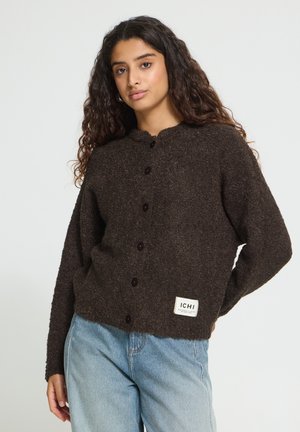 Jeune femme aux longs cheveux bouclés portant un cardigan marron foncé boutonné et un jean bleu clair sur un fond uni.