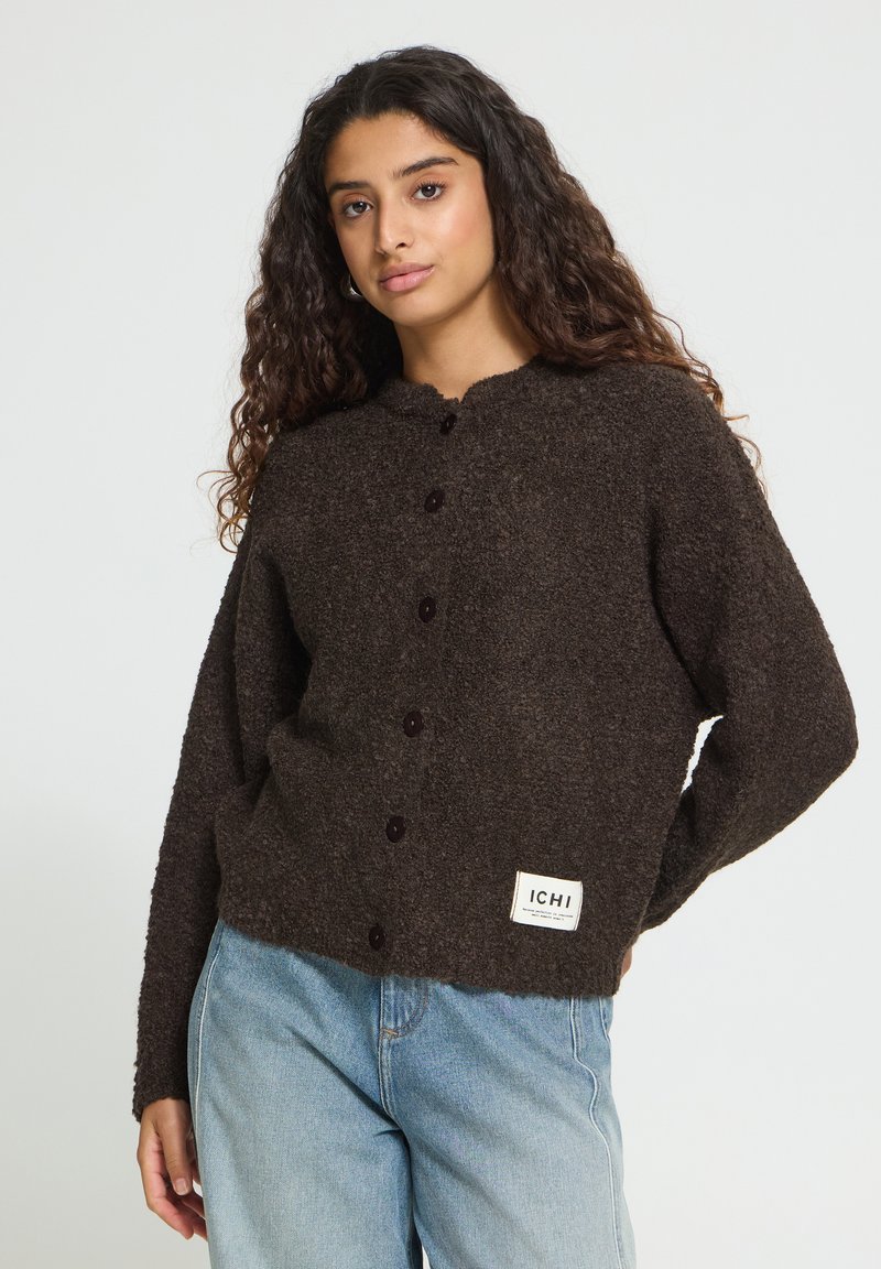 Jeune femme aux longs cheveux bouclés portant un cardigan marron foncé boutonné et un jean bleu clair sur un fond uni.