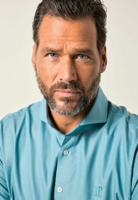 Středněvěký muž se šedomodrýma očima a vousy šedivě peprmintového vzhledu, oblečený v tyrkysové košili na knoflíky, hledící přímo před sebe s neutrálním výrazem.