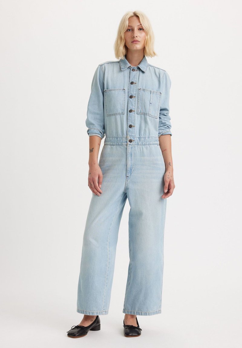 Jasnoniebieski kombinezon jeansowy z długimi rękawami, zapinany na guziki z przodu, z dwoma kieszeniami na piersi i szerokimi nogawkami. Gładka tekstura i luźny krój.