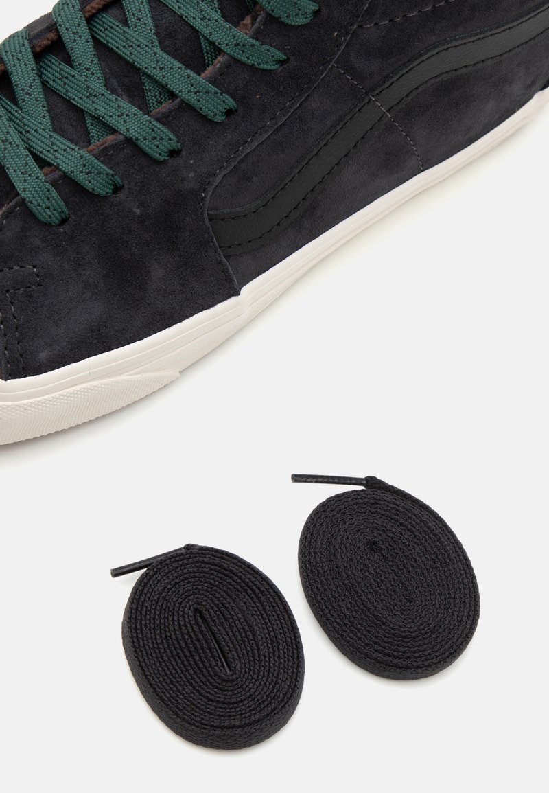 Zapatillas de ante gris oscuro con acentos en negro, suela de goma blanca y cordones verdes. Incluye dos cordones redondos negros.