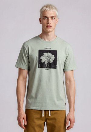 Giovane uomo con capelli biondi corti indossa una t-shirt verde chiaro con una grafica floreale in bianco e nero e la parola "unless." sul davanti.