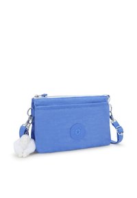 Blå nylon crossbody-väska med en texturerad yta, som har en rund logotyp, två zipfack och en fluffig vit charm fäst vid den.