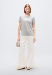 Γκρι βαμβακερό μπλουζάκι με λευκό καλλιγραφικό κείμενο "Boss", συνδυασμένο με φαρδιά λευκά παντελόνια. Το μοντέλο κρατά μια μπεζ τσάντα και φορά μπεζ παπούτσια.