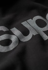 Tejido negro con un gran texto plateado y reflectante que dice "sup." La textura parece suave y ligeramente brillante.