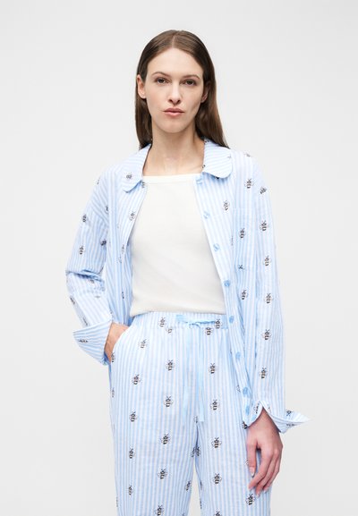 Kvinde iført lyseblåt og hvidt stribet pyjamassæt med bi-mønstre og en hvid top, stående mod en ensfarvet lys baggrund.