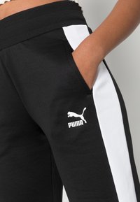 Pantalones deportivos negros con paneles laterales blancos, cinturilla acanalada y logotipo de PUMA impreso en blanco en la parte inferior de la pierna. Textura de tela suave.